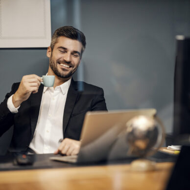 A cheerful executive sitting in his office and drinking coffee while smiling at the laptop.
