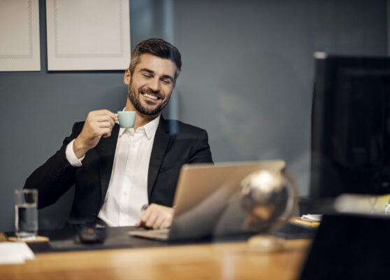 A cheerful executive sitting in his office and drinking coffee while smiling at the laptop.